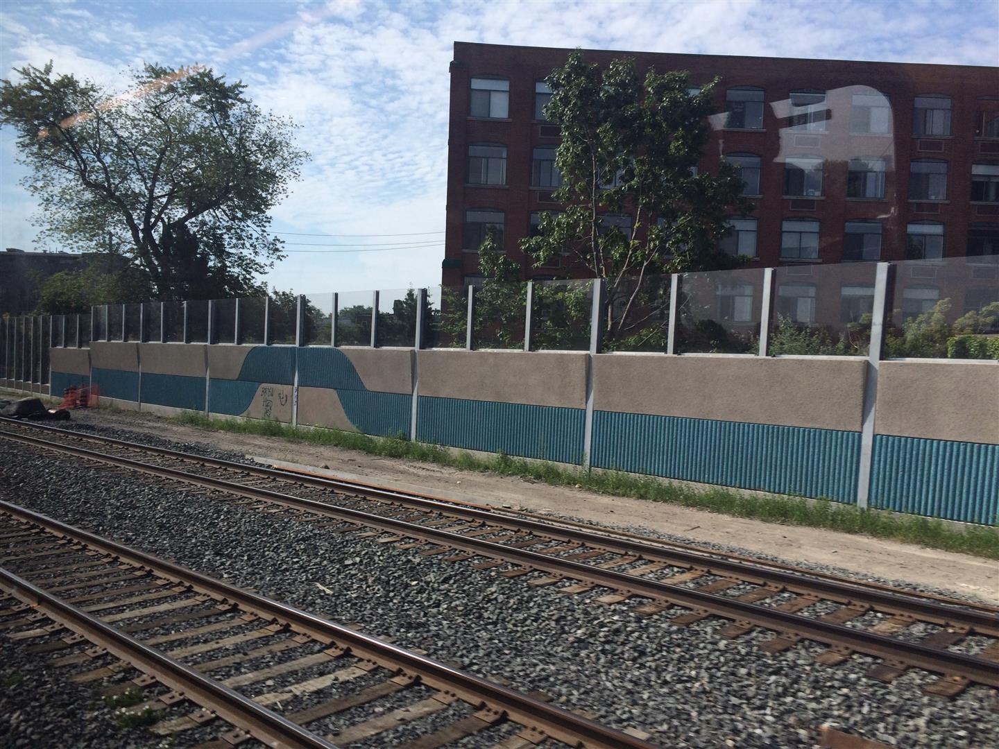 Noise barrier panels featuring a custom "ribbon" pattern in blue topped with Transparent Acrylite Soundstop sheets along Metrolinx's Union Pearson Express rail line in Toronto, ON.